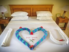 Una cama con sábanas blancas, un arreglo de toallas en forma de corazón que deletrea "Mr & Mrs" con flores rojas, y una toalla enrollada al lado.