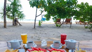 Mesa de desayuno con diversos alimentos y bebidas, con vista a una playa que tiene sillas de descanso y palmeras.