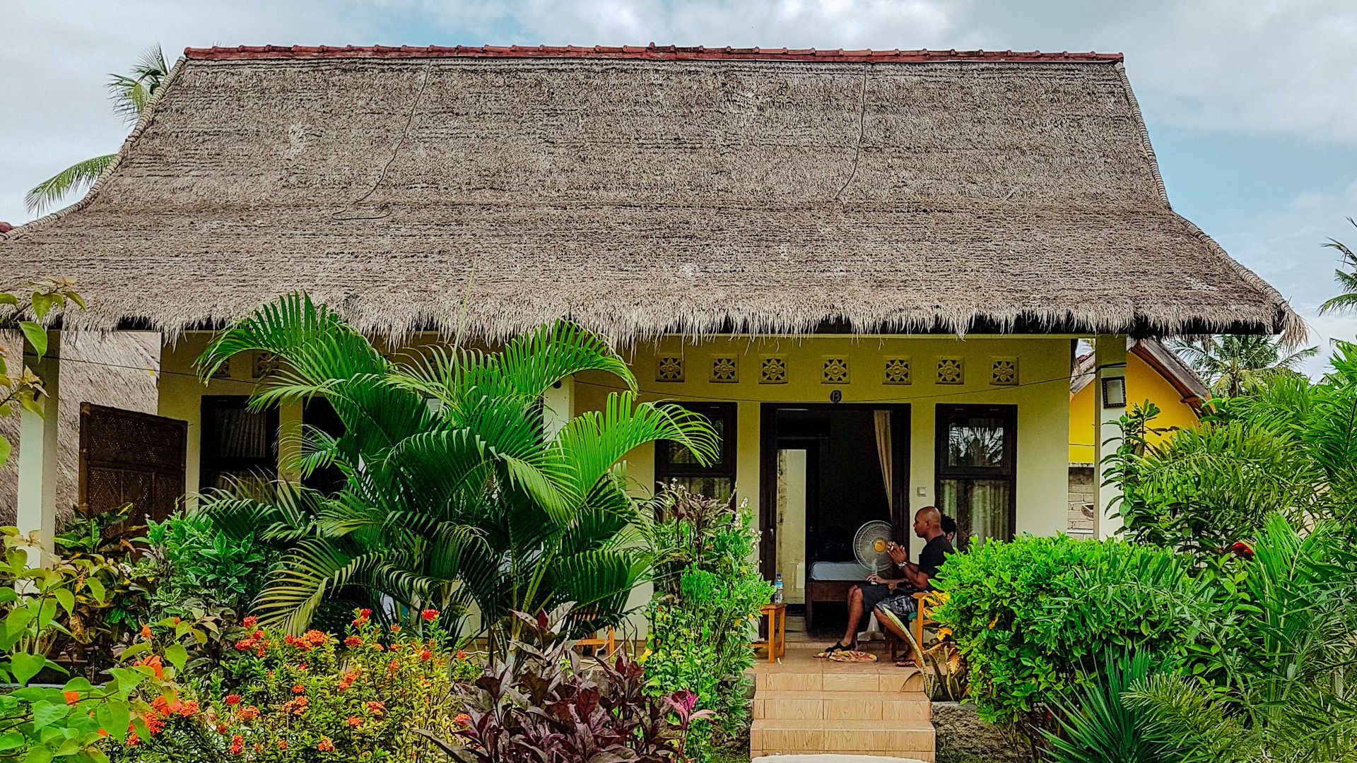 A thatched-roof house with a porch, surrounded by lush tropical plants and flowers.