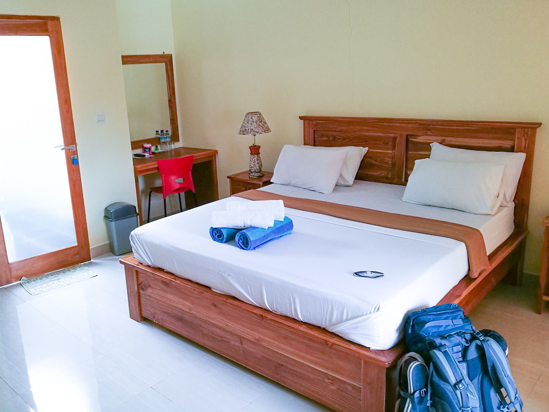 A double bed with white linens, a wooden headboard, and a blue towel on the bed. There is a wooden nightstand with a lamp, a red chair, and a desk with a computer and toiletries. A blue backpack is on the floor next to the bed.