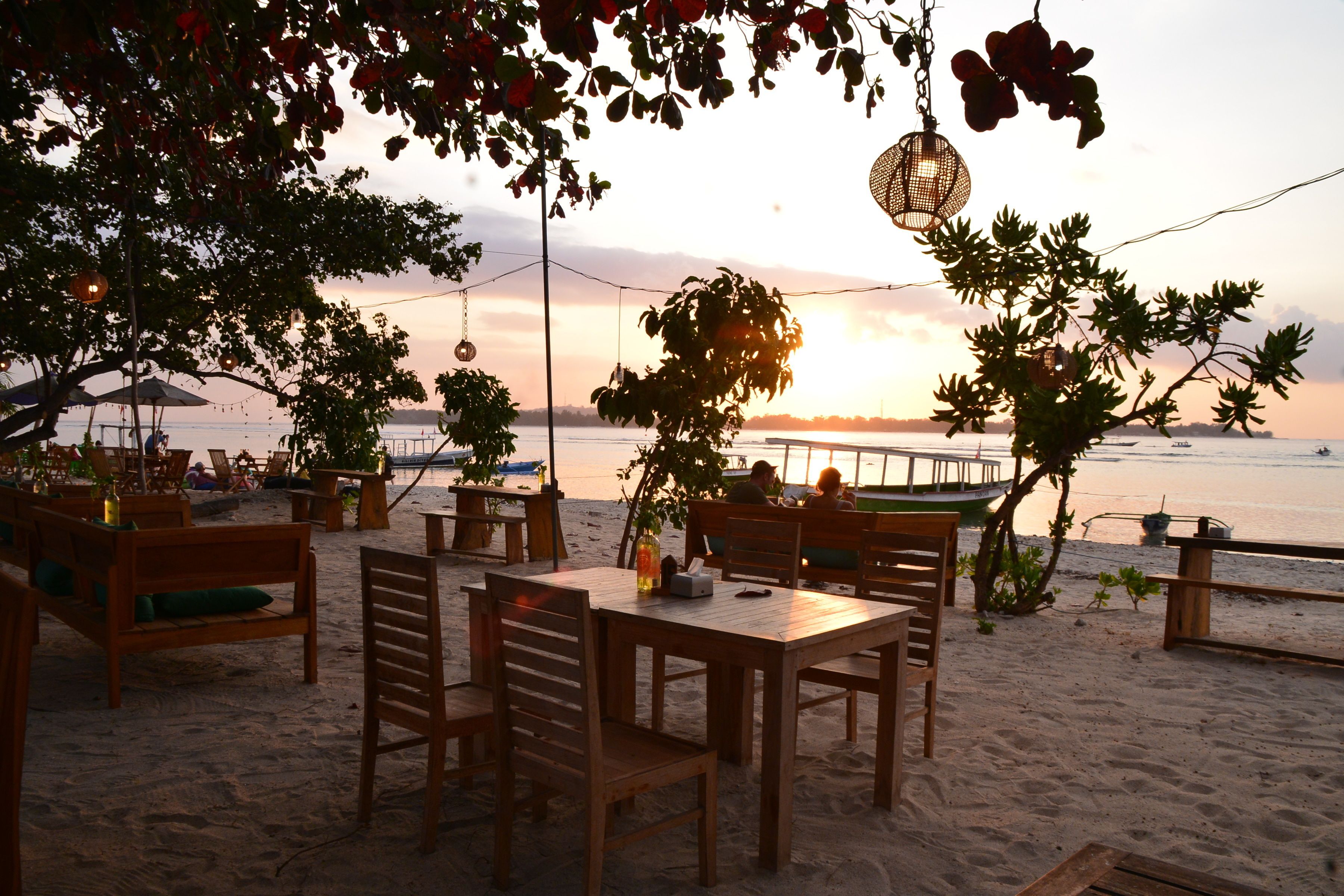 Un restaurante junto a la playa con mesas y sillas de madera, terraza y vistas a la puesta de sol sobre el agua.