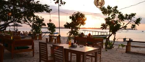 Un restaurante junto a la playa con mesas y sillas de madera, terraza y vistas a la puesta de sol sobre el agua.