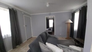 A living room with a grey sofa, a lamp, and a view of a bedroom through an open door. - Kalgan River Chalets and Caravan Park (Kalgan)