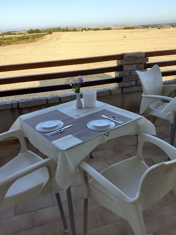 A table set for two with white plastic chairs, a vase with flowers, and a view of a field and beach. - Il Tramonto (Giba)