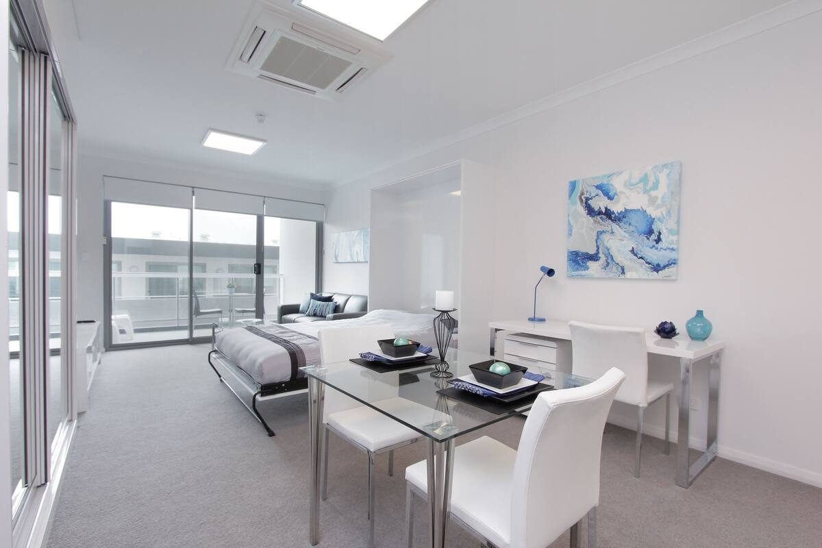 A modern hotel room with a glass dining table, white chairs, a bed with a gray and white striped blanket, a desk with a lamp, and a large abstract painting on the wall.