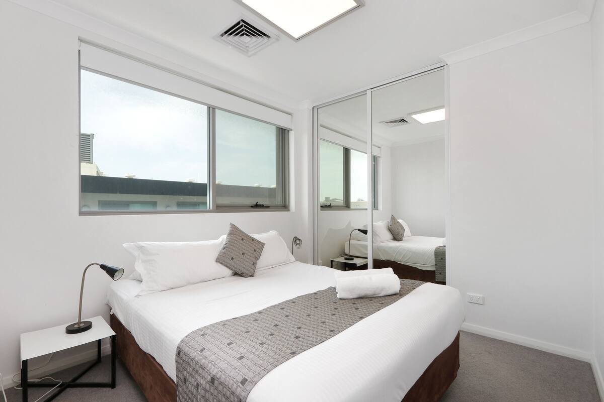 A modern hotel room with a large bed, a bedside table, a mirror, and a window with a city view.