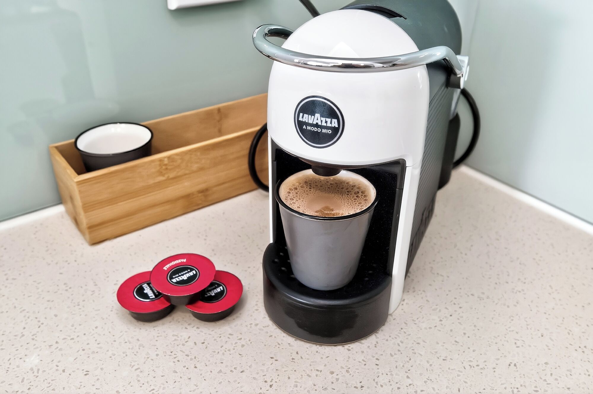 Een koffieapparaat met kopjeshouder, een houten doos met een kopje, en twee rode koffiekapsules.
