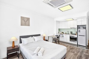 A hotel room with a bed, bedside lamps, a painting, a kitchen area with appliances, and a dining table.