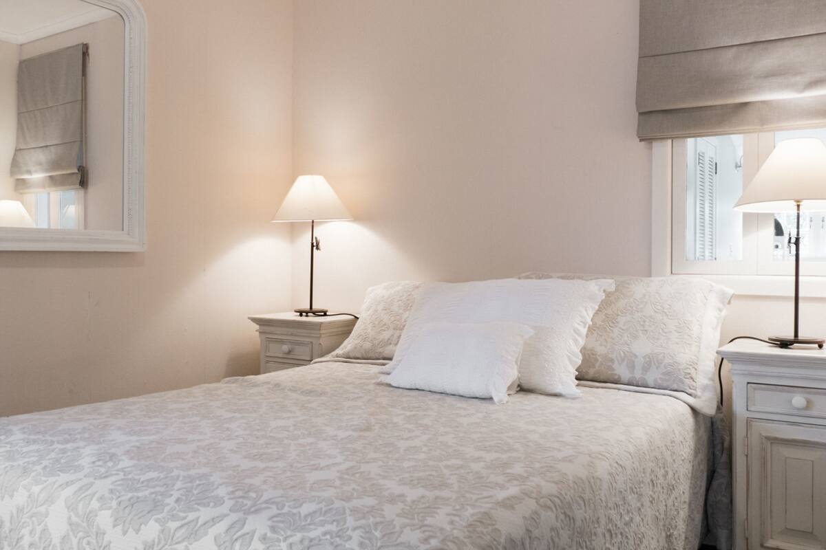 Un dormitorio con cama, dos mesitas de noche, una lámpara, una ventana con persiana y un espejo en la pared.