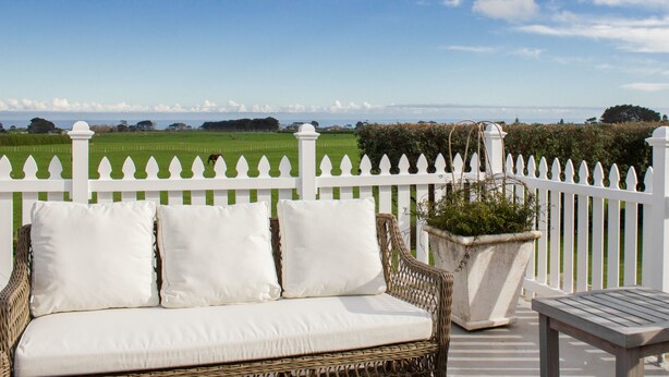 A wicker sofa with white cushions on an outdoor deck, overlooking a green field and a white picket fence. - Goodin Country Garden (Warea)