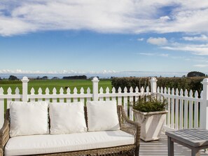A wicker sofa with white cushions on an outdoor deck, overlooking a green field and a white picket fence. - Goodin Country Garden (Warea)