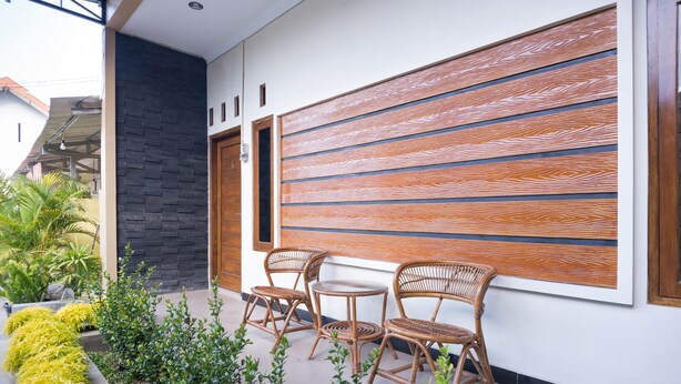 A modern outdoor seating area with wicker chairs and a small table, surrounded by neatly trimmed plants and a decorative wall panel. - Hotel O Al Abror Homestay (Yogyakarta)