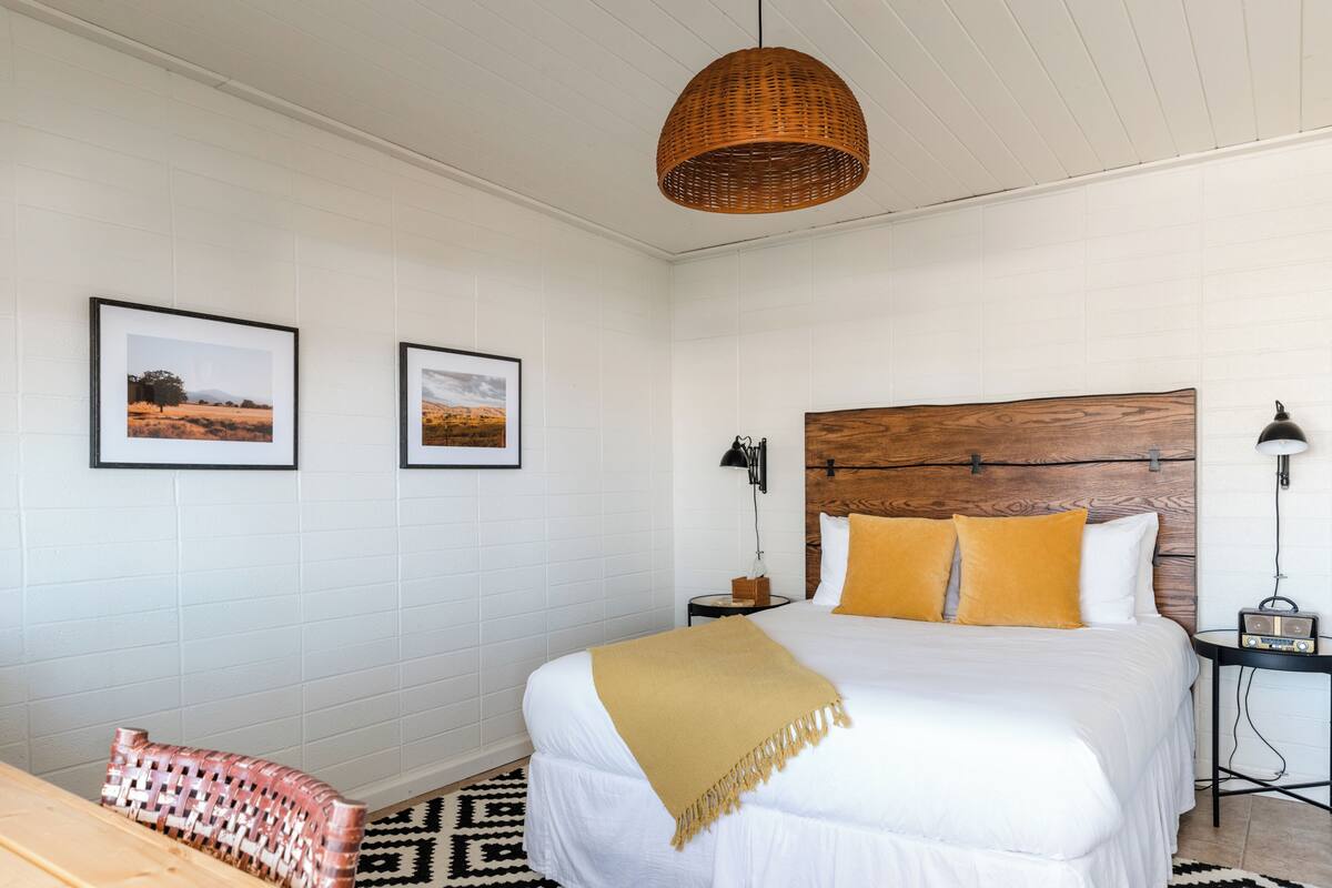 A neatly arranged bedroom with a large bed, a bedside table with a lamp, and two framed pictures on the wall.
