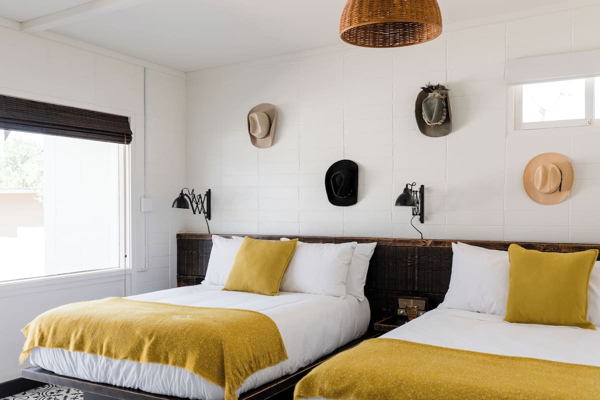 A hotel room with two beds, a large window with blinds, and several hats hanging on the wall.