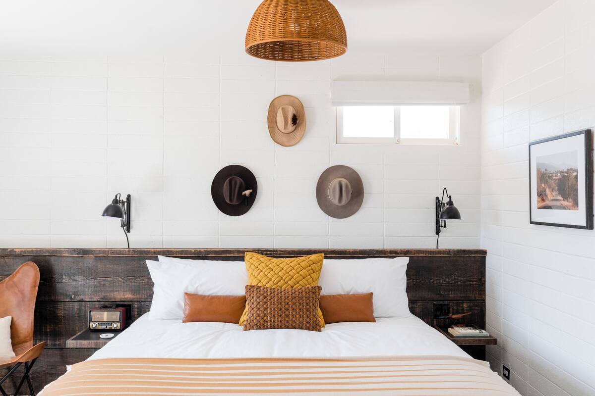 A hotel room with a bed, a chair, a nightstand, a radio, and a wall with hats and a picture.