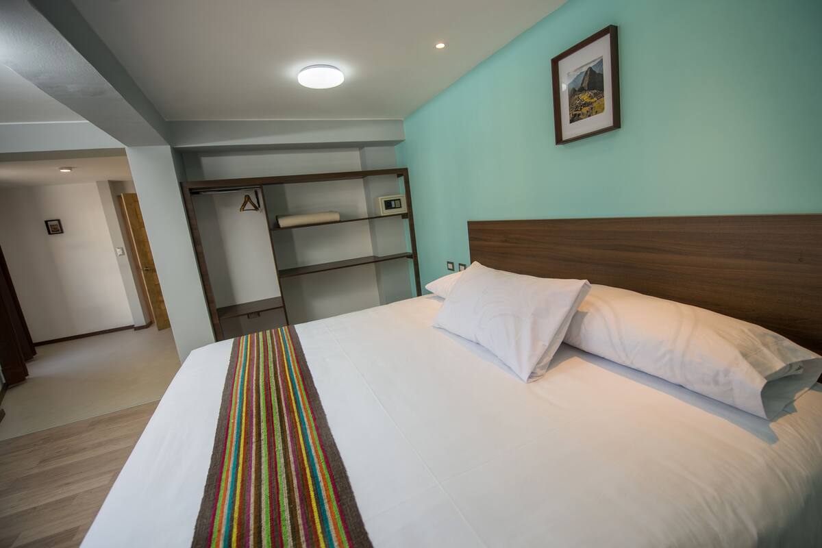 A hotel room with a bed, a colorful striped blanket, a wooden headboard, a shelf with a clock, and a framed picture on the wall.