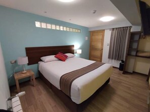 A hotel room with a large bed, a wooden headboard, a nightstand with a lamp, a window with blinds, and a door with a curtain.