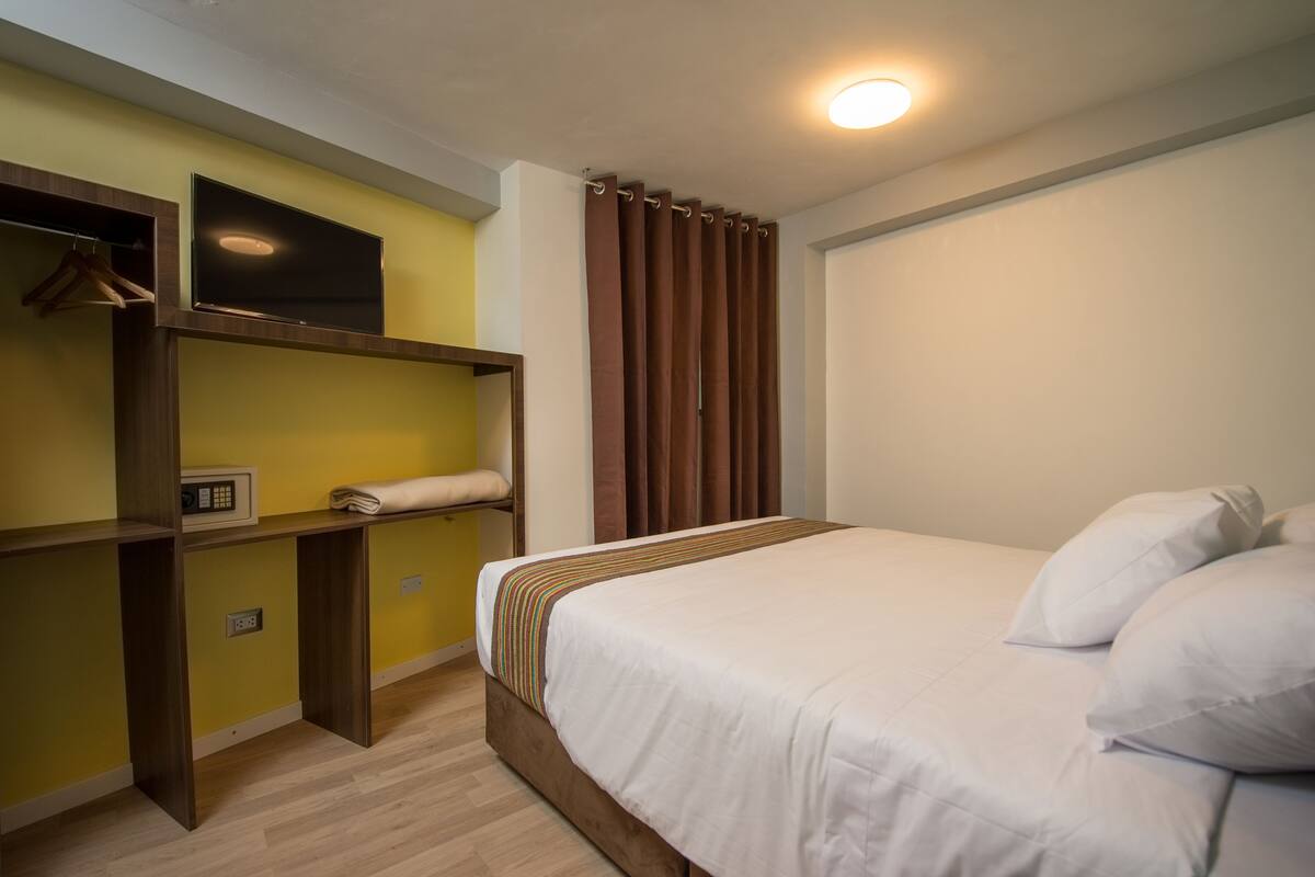 A hotel room with a bed, a TV mounted on a wall, a desk with a chair, and a window with curtains.