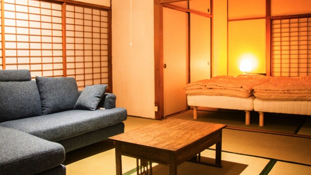A traditional Japanese room with tatami flooring, a low wooden table, a sofa, and sliding doors leading to a bed area. - Engawa (Tonosho)