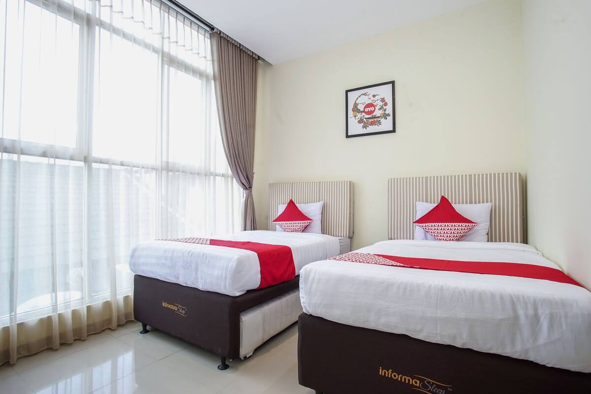 A hotel room with two beds, each with white bedding and red accents, a large window with sheer curtains, and a framed picture on the wall.