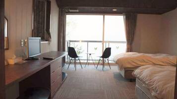 A hotel room with a desk, two chairs, a bed, and a large window.