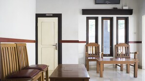 Een kamer met een houten bank, een tafel en twee stoelen. Er is een deur met daarop 'TOILET' en een televisie aan de muur bevestigd.