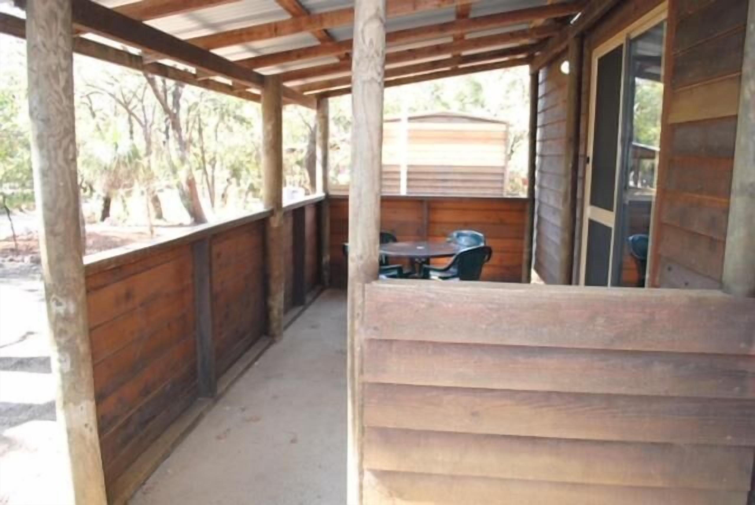 A covered outdoor area with a wooden deck, a table, and chairs, surrounded by wooden walls and a view of trees.