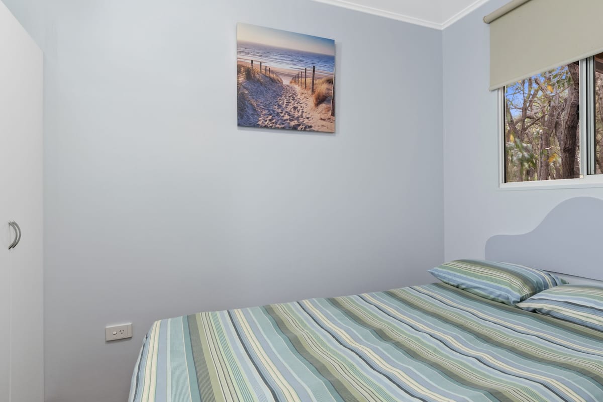 Um quarto com uma cama, uma pintura de uma praia e uma janela com vista para árvores.