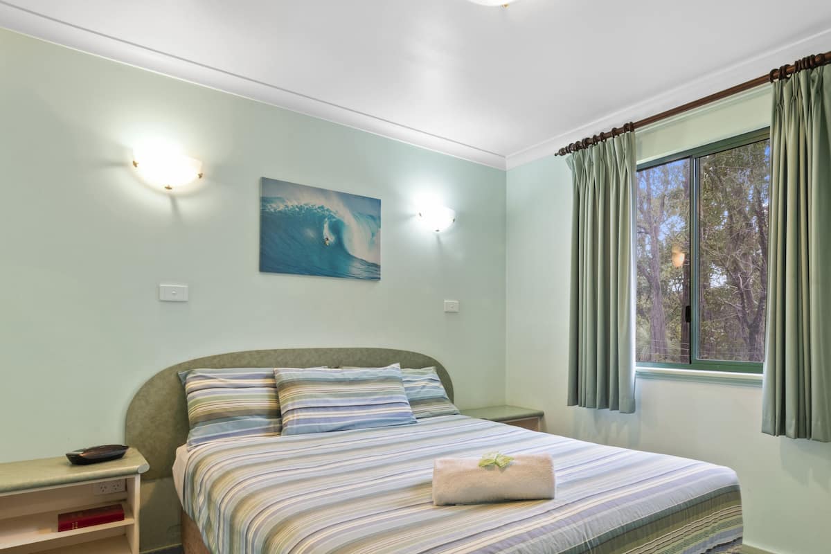 Quarto com cama, mesas de cabeceira, um criado-mudo, janela com cortinas e luminária fixada na parede.