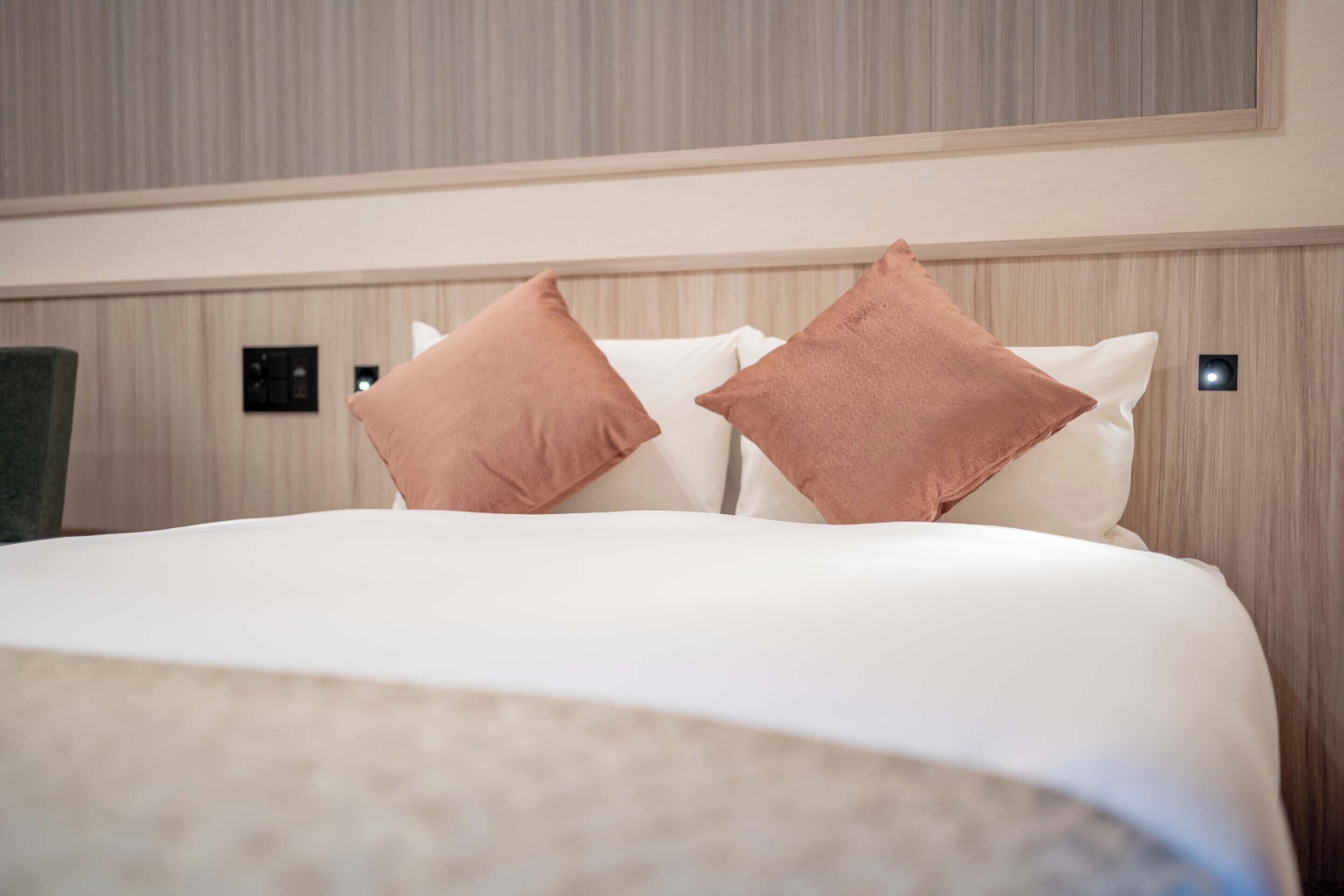 A hotel room with a bed, two pillows, a headboard, and a wall-mounted light.