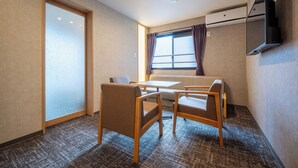 Hotellihuoneessa on sohva, kaksi tuolia, pieni pöytä, seinälle asennettu televisio ja ikkuna, jossa on verhot.