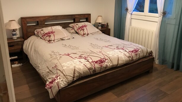 Een slaapkamer met een houten bed, bloemenbeddengoed, bedlampen, een raam met gordijnen en een radiator.