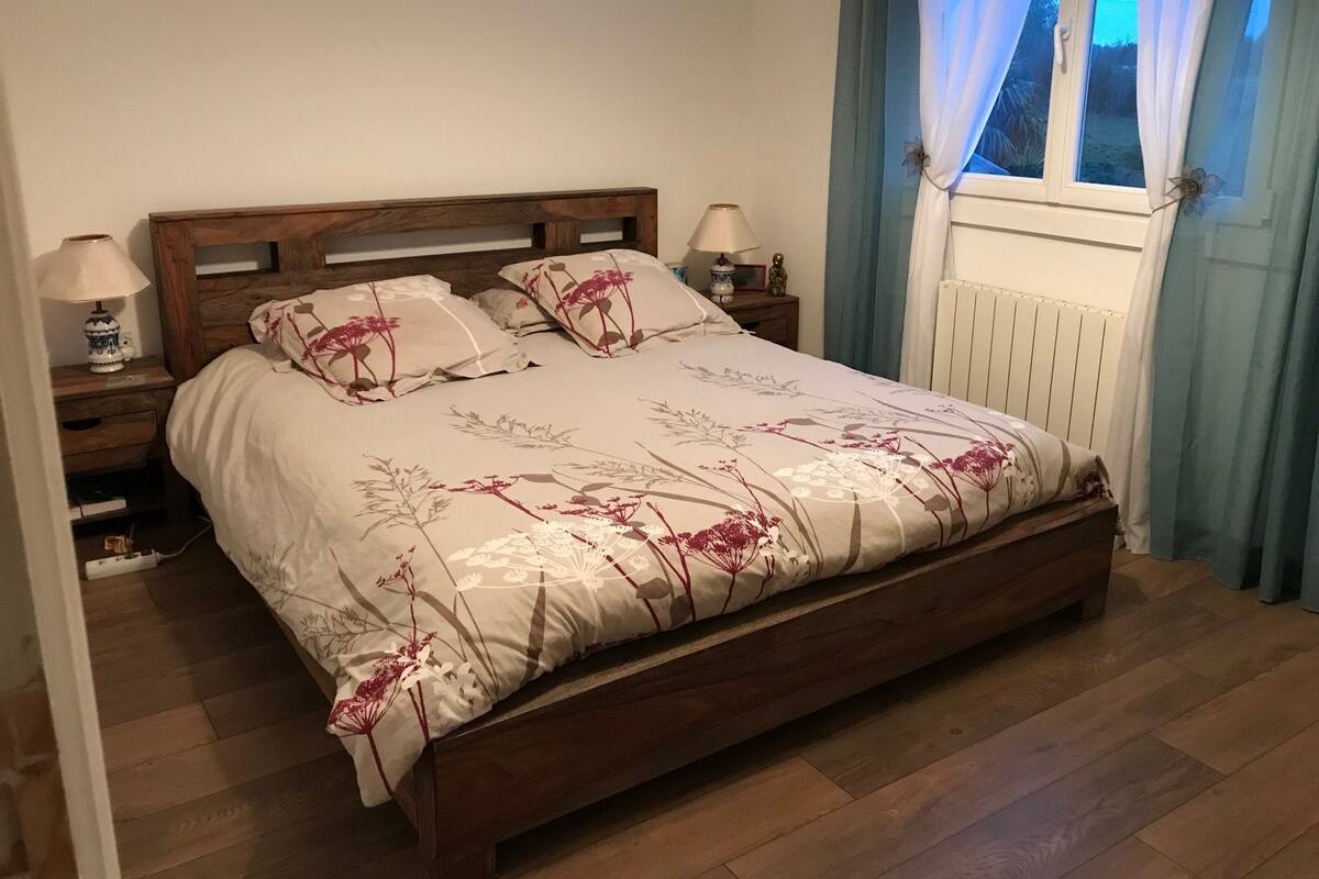 Een slaapkamer met een houten bed, bloemenbeddengoed, bedlampen, een raam met gordijnen en een radiator.