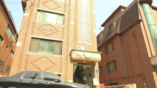 Un edificio de varios pisos con fachada de ladrillo, un coche estacionado adelante y una entrada visible con un cartel.