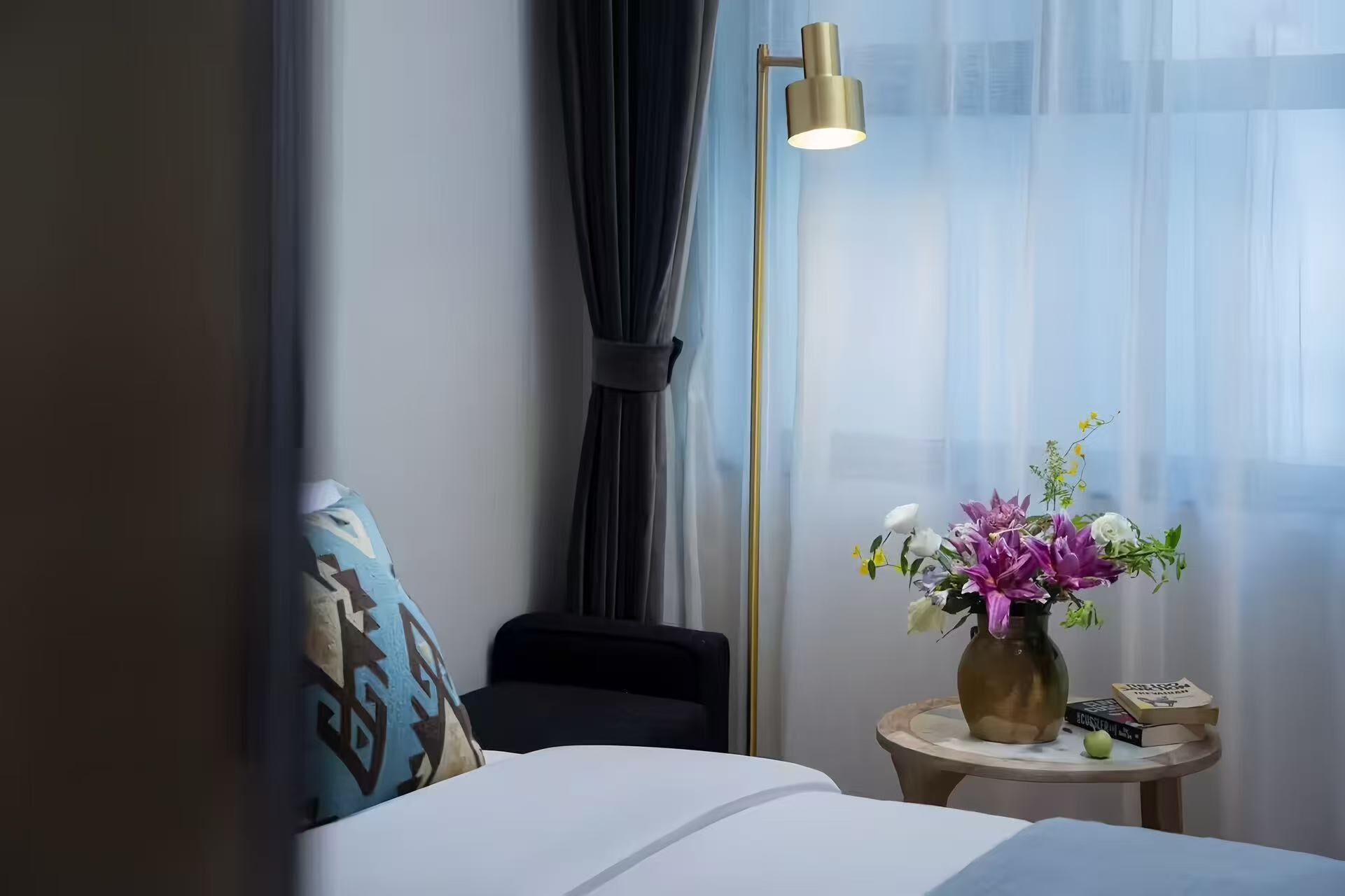 Una habitación de hotel con una cama, una mesita de noche con un jarrón de flores, una lámpara de pie y vistas al exterior a través de cortinas translúcidas.