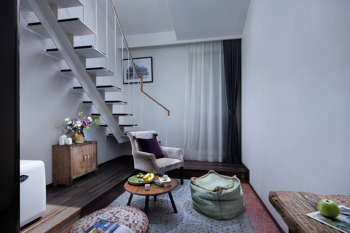Een moderne woonkamer met een trap, een comfortabele fauteuil, een ronde salontafel met fruit en een vaas, en een groene kruikzak.