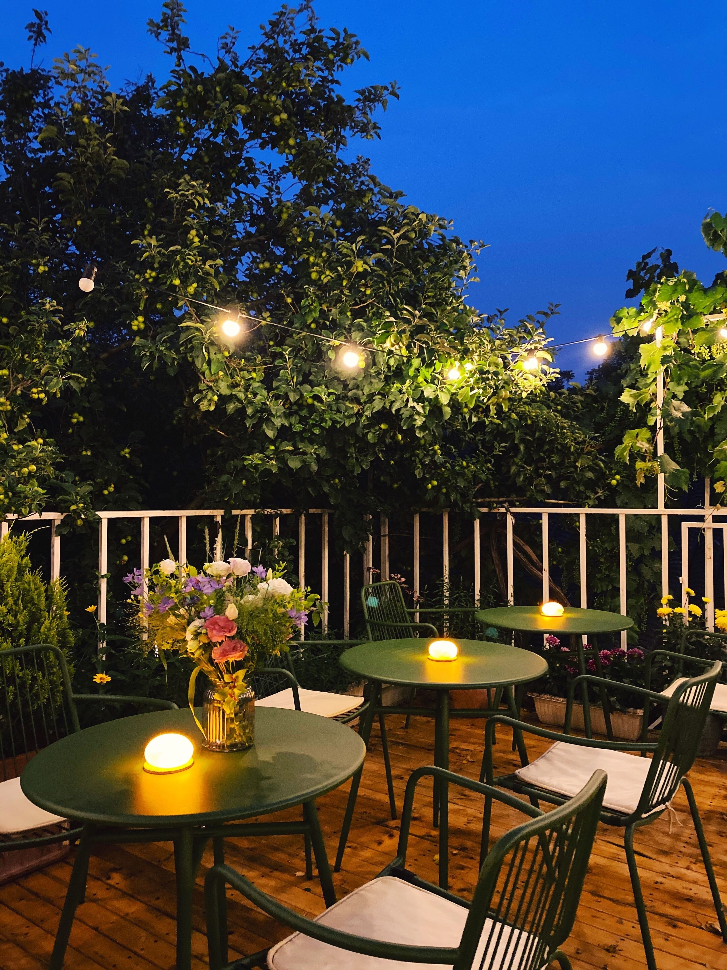 An outdoor seating area with round tables and chairs, surrounded by greenery and a wooden fence.