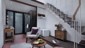 Un loft spacieux avec un plafond haut, des poutres apparentes, un escalier avec une rampe en bois, un coin salon avec une table et un fauteuil, et un petit meuble sur lequel est posé un vase de fleurs.