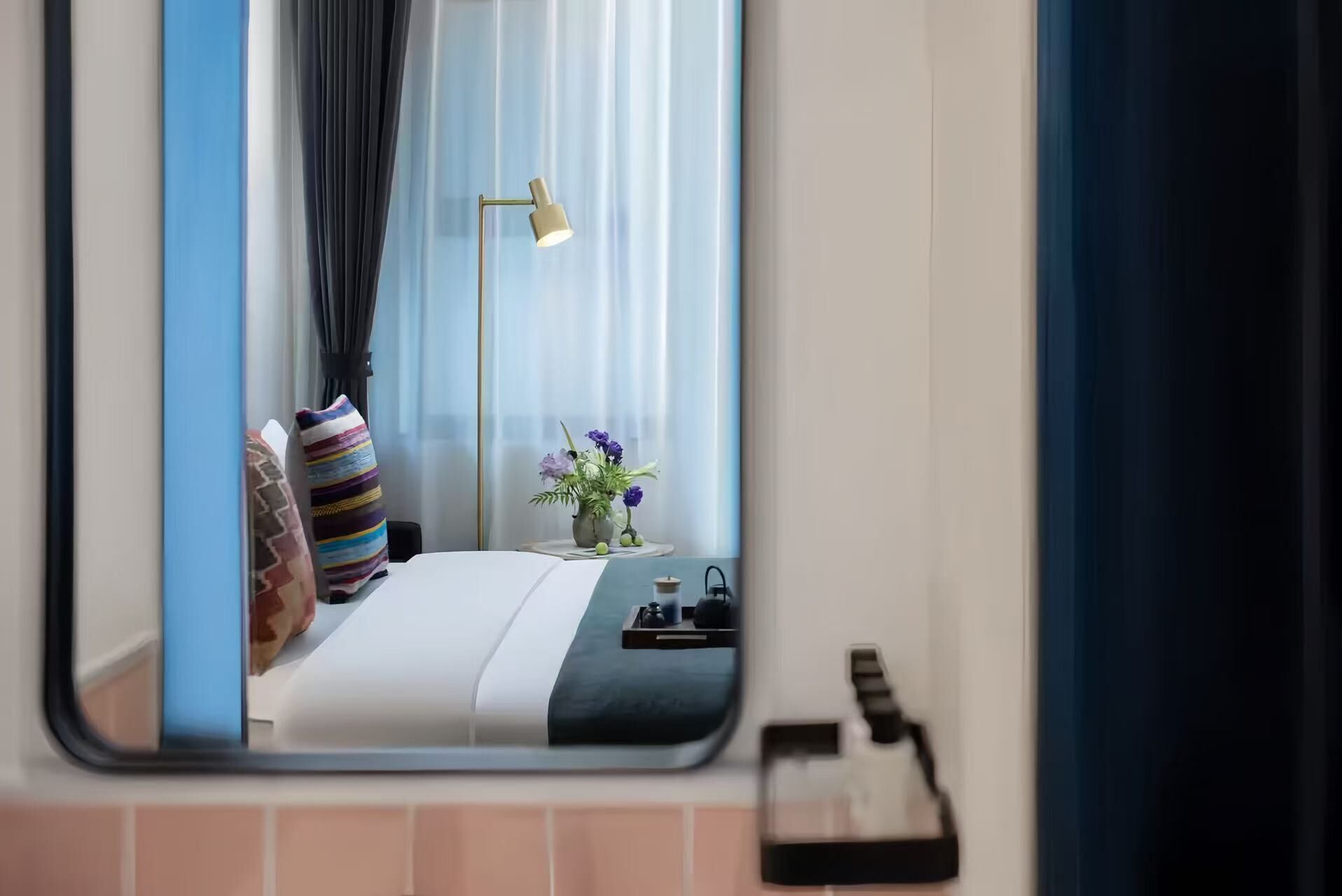 A hotel room with a bed, a bedside table with a lamp, a vase with flowers, and a window with curtains.