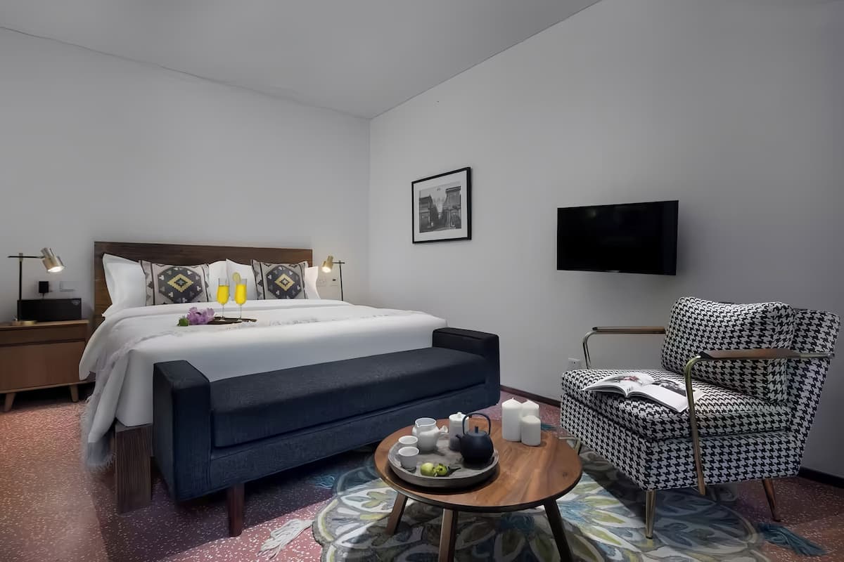 Een moderne hotelkamer met een bed, nachtkastje, lamp, televisie en een fauteuil met een hondentandpatroon.