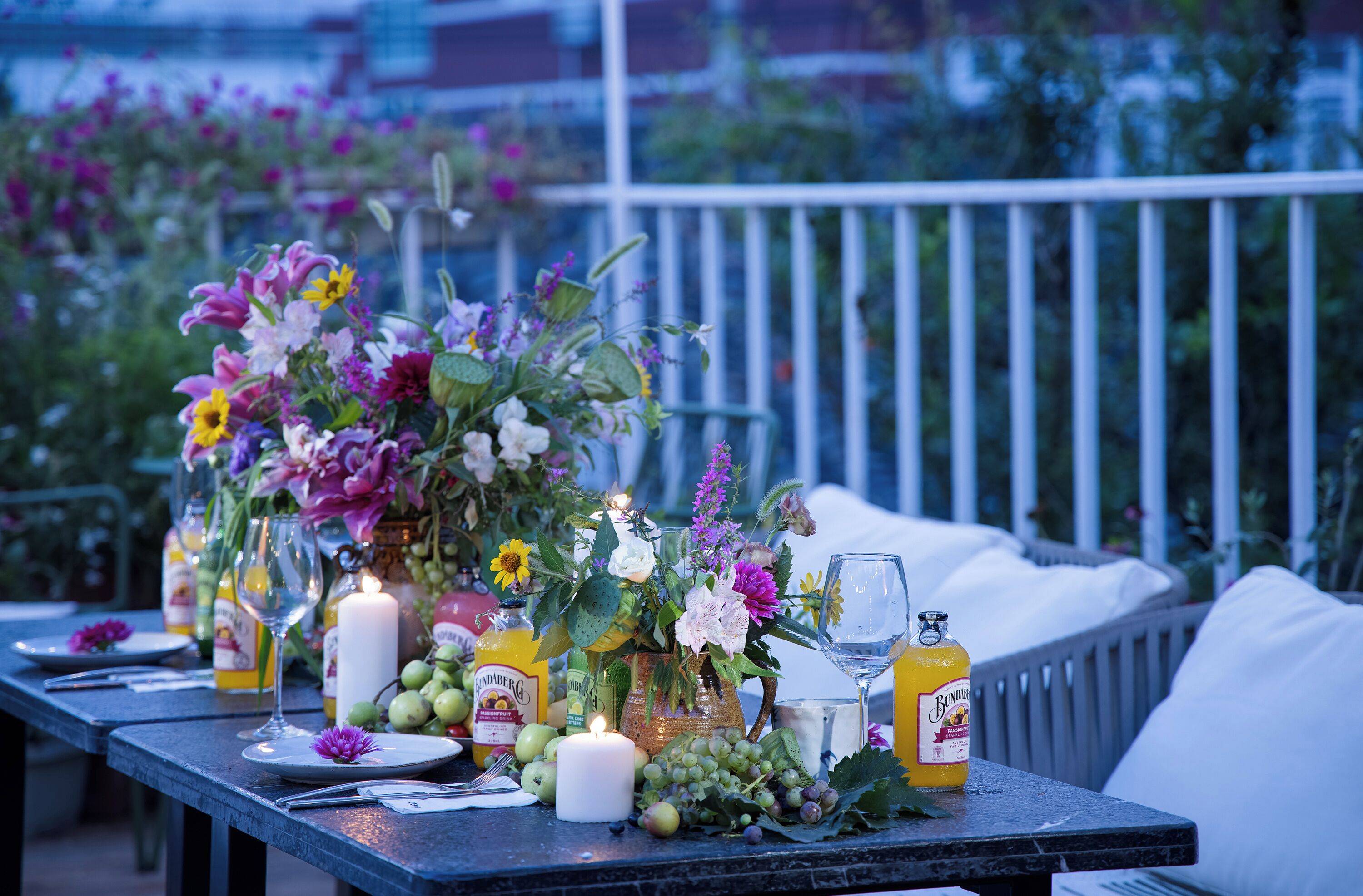 Una mesa adornada con una vela, copas de vino y una botella de jugo, rodeada de flores y follaje.