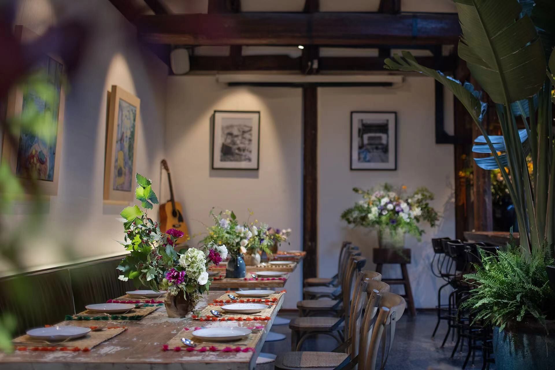 A long dining table with chairs, set for a meal, in a room with framed pictures on the walls and a guitar in the background.