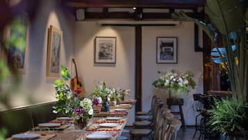Una larga mesa de comedor con sillas, dispuesta para una comida, en una habitación con cuadros en las paredes y una guitarra al fondo.