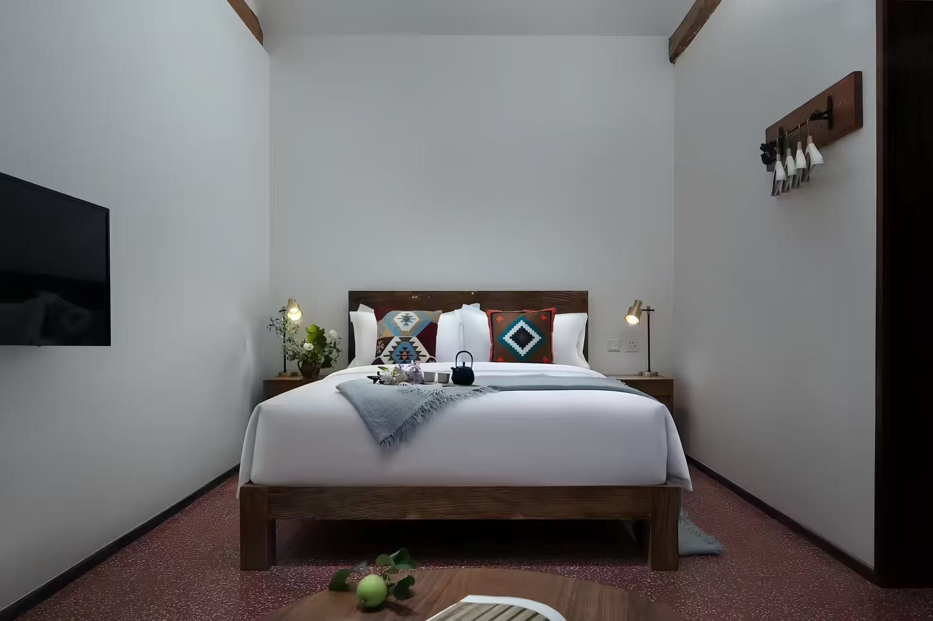A neatly arranged bedroom with a large bed, bedside tables, a wall-mounted TV, and a small table with a vase and teapot.