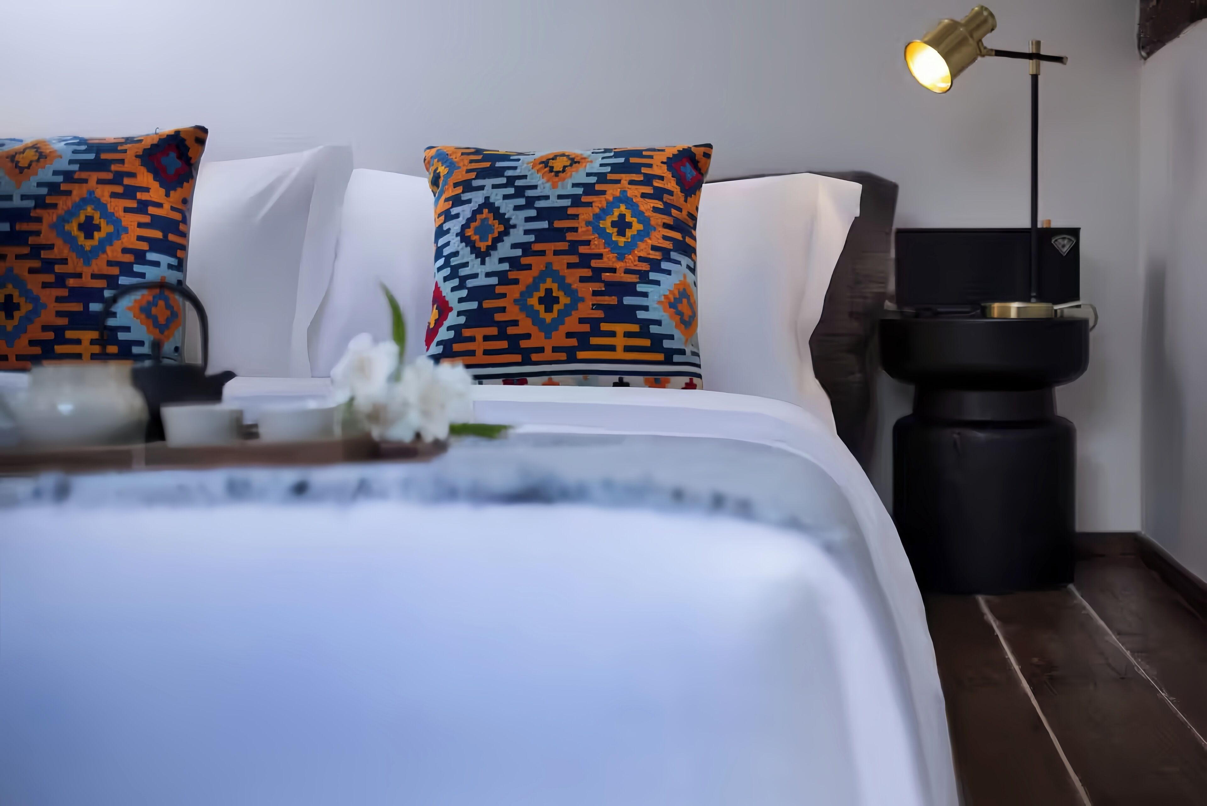 Una cama bien hecha con almohadones estampados, una mesita de noche con una lámpara y un pequeño cubo de basura negro.