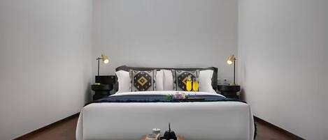 Un dormitorio con una cama grande, dos mesitas de noche con lámparas, un banco de madera y una pequeña mesa con objetos decorativos.