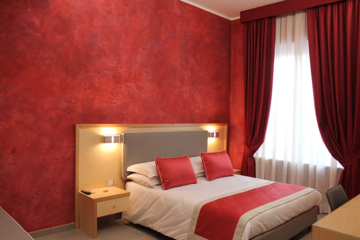A hotel room with a red textured wall, a bed with white bedding and red pillows, a bedside table with a lamp, and a window with red curtains.