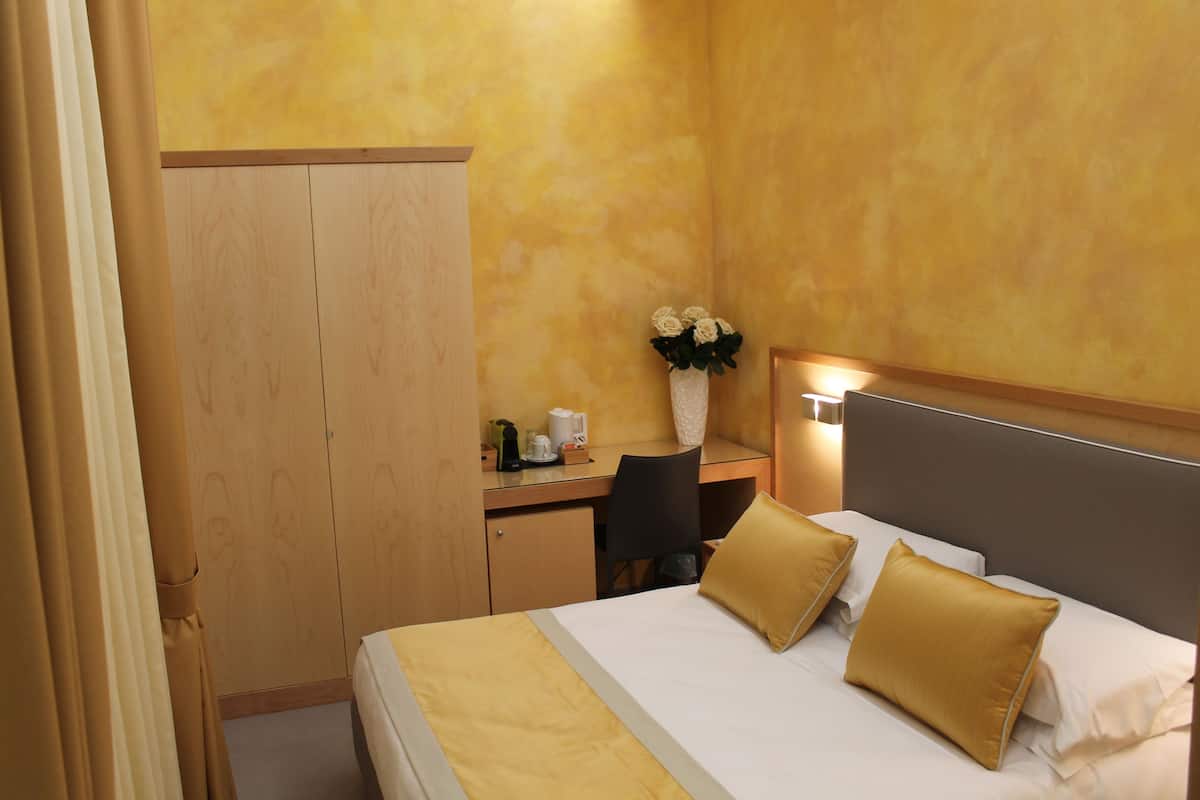 A hotel room with a bed, a desk, a chair, a wardrobe, and a vase with flowers.