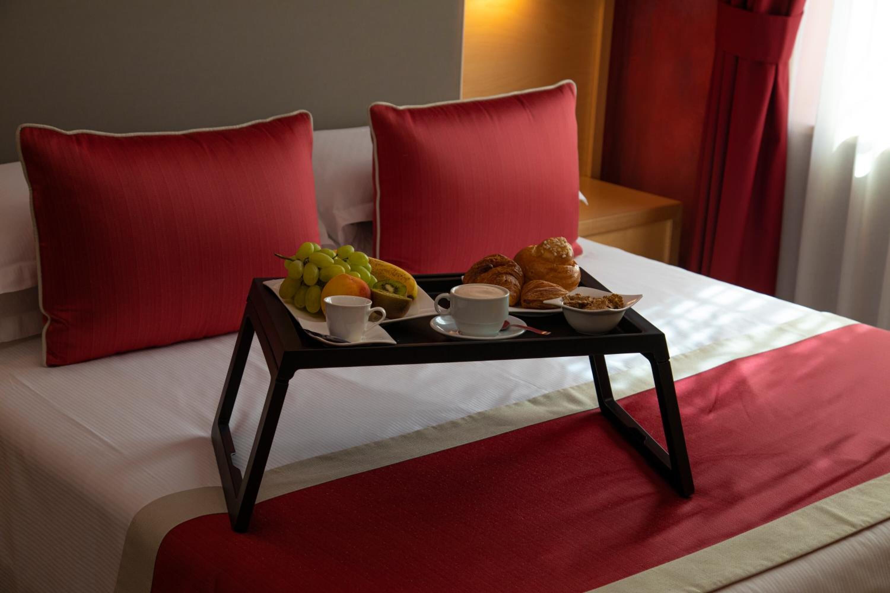 A neatly made bed with red pillows, a tray with breakfast including fruits, pastries, and a cup of coffee.
