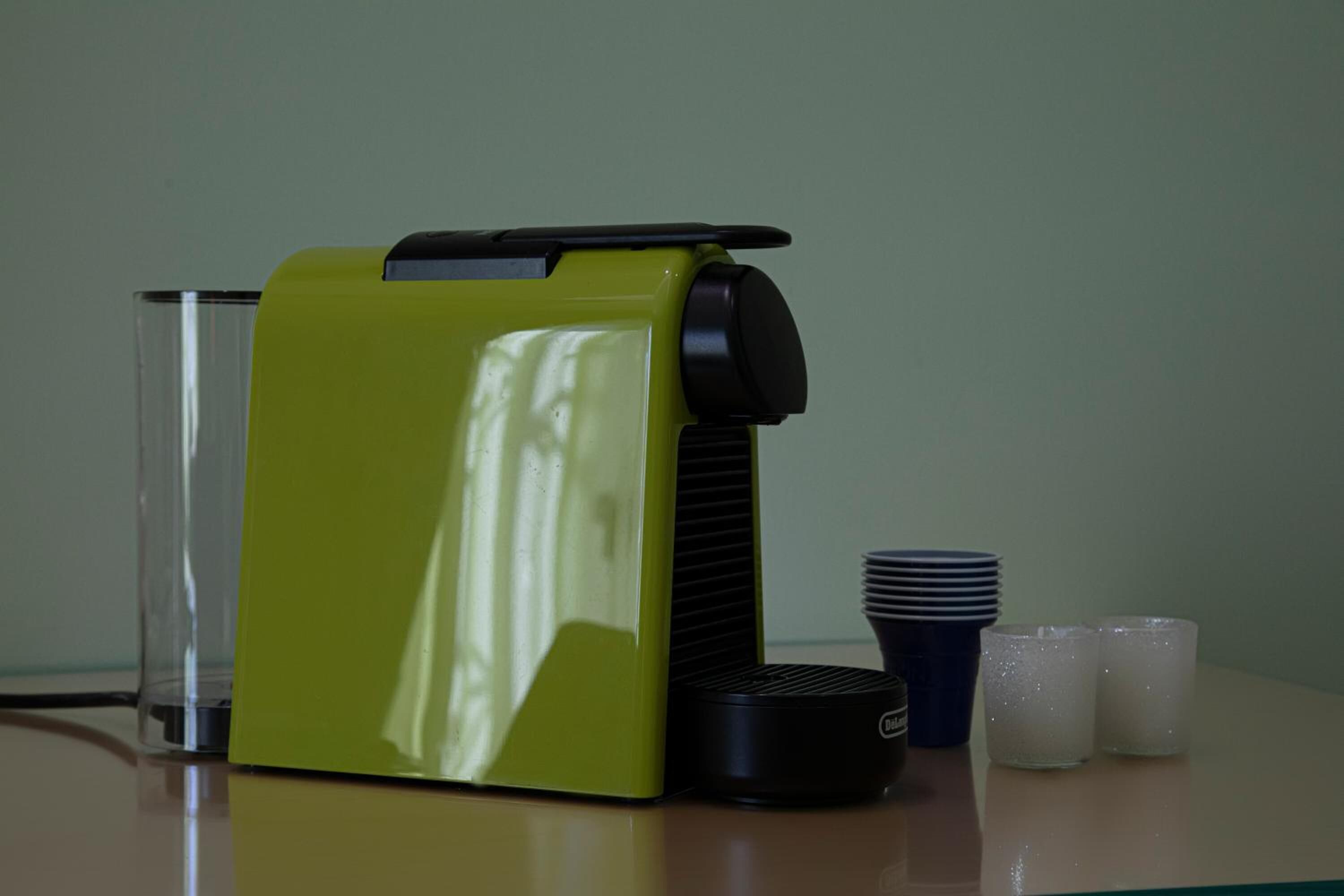 A green coffee machine with a black top, a clear glass, and a stack of blue and white cups on a table.